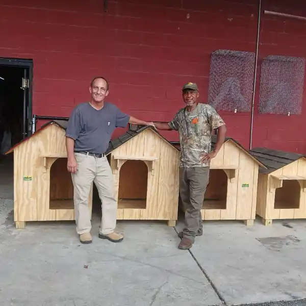 Two men from the shop standing in front of a red barn wall lined with finished dog houses